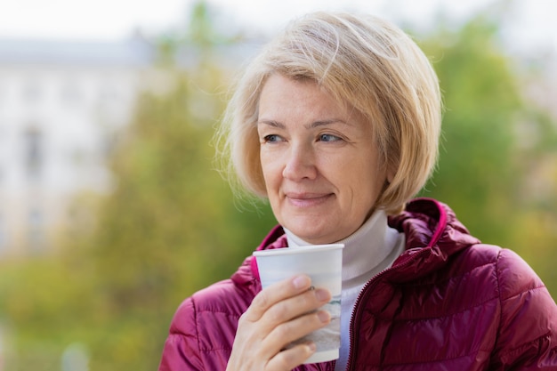 Elegant senior adult woman outdoors with coffee, looking to the side and smiling