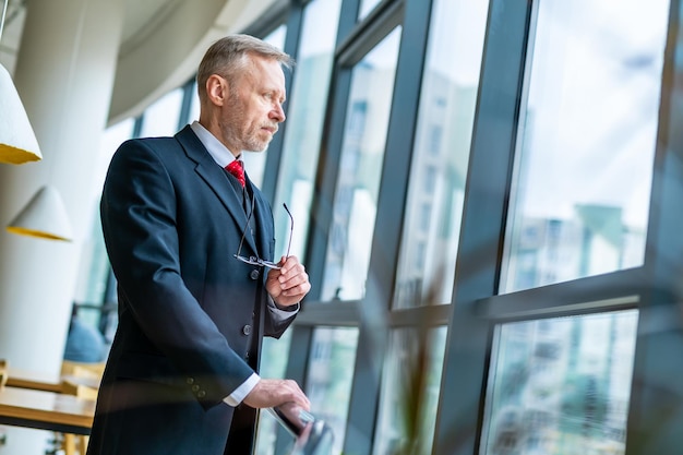 Elegant senior man in dark suit and red tie is looking to the panoramic window. View from below.