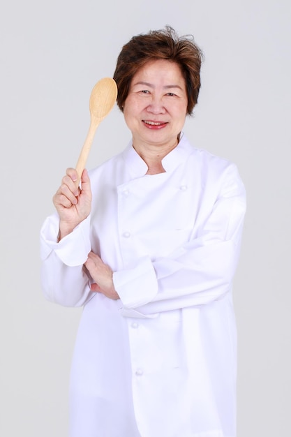 레스토랑 주방에서 팔짱을 끼고 나무 숟가락과 포크를 들고 자신 있게 서 있는 전문 요리사로서의 우아한 고위 여성. 은퇴하신 분들의 건강한 요리를 위한 요리의 달인 이모님.