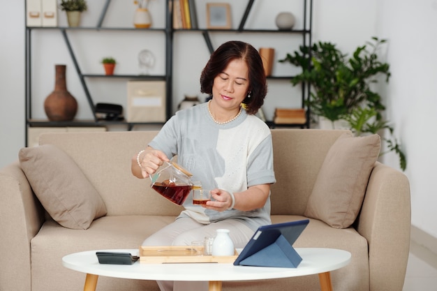 Elegante donna anziana seduta sul divano di casa e versandosi una tazza di caffè