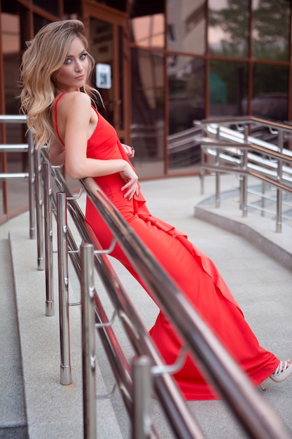 Elegant sexy blue-eyed girl in an evening red dress posing leaning on the railing of the street stairs