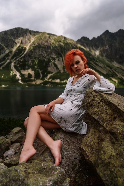 Elegant sexy lady with red hair in white sits on the stone in mountains and relaxes under sun rays