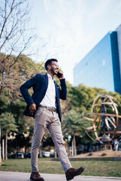 Elegant smiling black businessman walking in a park while using his smartphone