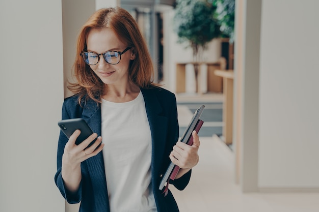 Elegante donna sorridente impiegata in occhiali che utilizza un moderno telefono cellulare nello spazio di coworking