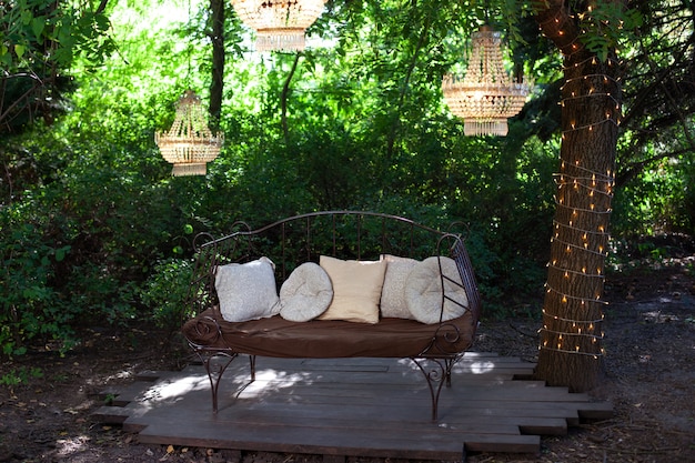 Элегантный диван в саду, наружная декоративная композиция с тремя люстрами. Красивый элегантный декор для свадебной церемонии в прекрасном саду. Беседка для отдыха на природе. Романтическая беседка
