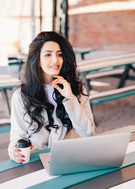 우아한 성공적인 여성 프리랜서는 공식적으로 원격 작업을 위해 일반 랩톱 컴퓨터를 사용하여 옷을 입고 꿈꾸는 표정을 가진 맛있는 커피를 마시는 야외 카페에서 휴식을 취합니다. 라이프 스타일 컨셉