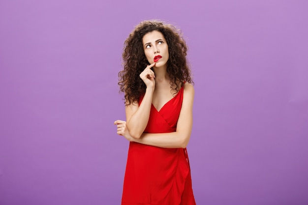 Elegant thoughtful woman in trendy red dress with curly
hairstyle and party makeup touching lip looking at upper right
corner thinking how make impression during conversation over purple
background.