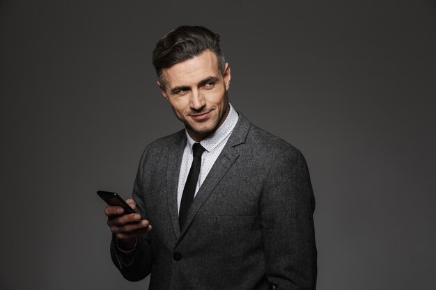 Elegant trendy man dressed in business costume holding smartphone and looking aside with smile, isolated over gray wall