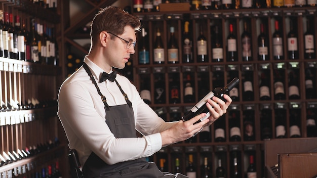 Elegant wine seller or cavist, offers a bottle of red wine, a man tells about the wine varieties, wine shelves in the background