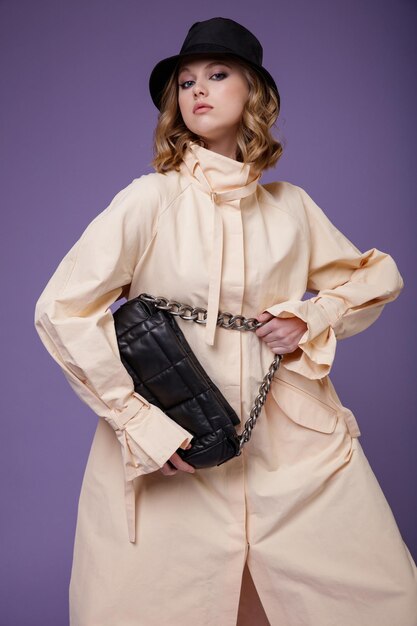 Elegant woman in beige cream trench coat black hat boots handbag on purple lilac background