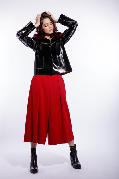 Elegant woman in black leather jacket red trousers pants posing over white background