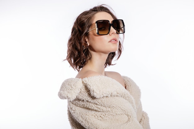 Элегантная женщина в кремово-бежевых шубах и стильных солнцезащитных очках позирует на белом фоне