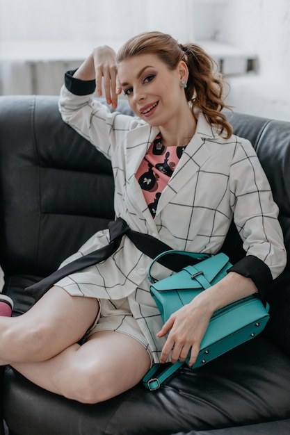 Elegant woman in a fashionable checkered suit pink blouse holding a turquoise backpack and posing sitting on the sofa