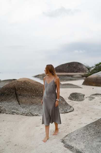 Donna elegante in abito di seta grigio in posa sulla spiaggia solitaria con tempo nuvoloso