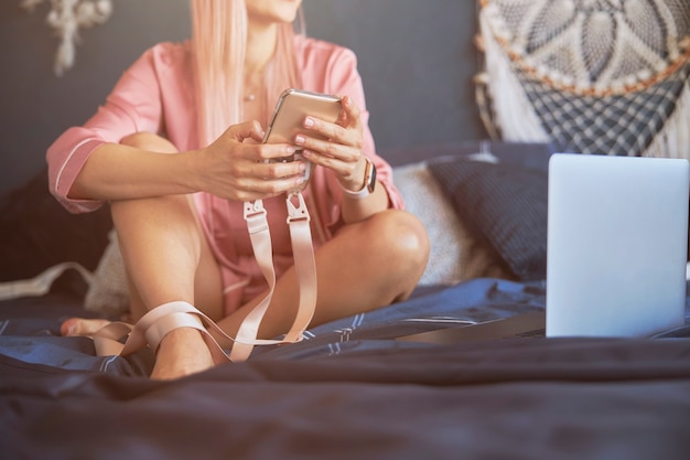 Elegant woman holds mobile phone sitting by modern laptop on bed with dark blue linen