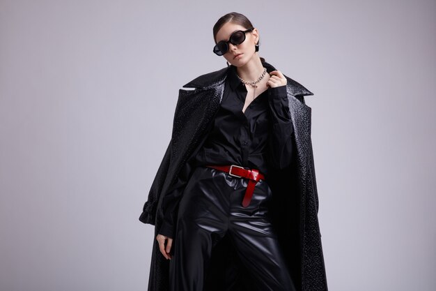 Elegant woman in leather coat pants sunglasses accessories shirt red belt on gray background