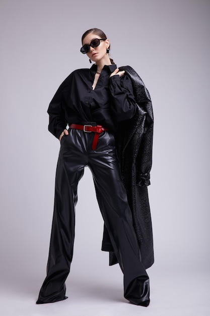灰色の背景に革のコートパンツサングラスアクセサリーシャツ赤帯のエレガントな女性