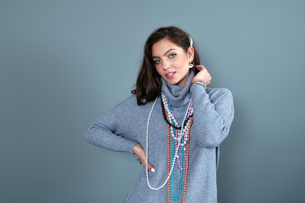 Elegant woman in a long gray sweater has fun with many bead chains and jewelry isolated on gray