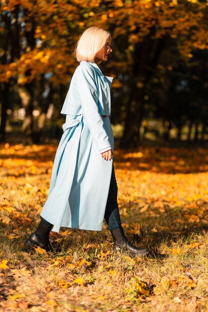 Elegant woman model in fashion blue coat with boots walks in the park with yellow fall foliage on a sunny autumn day
