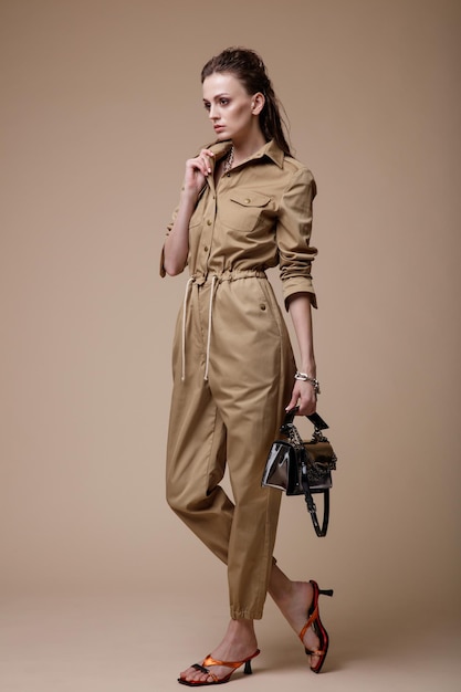 Elegant woman in pretty beige sand jumpsuit accessories handbag posing on brown background