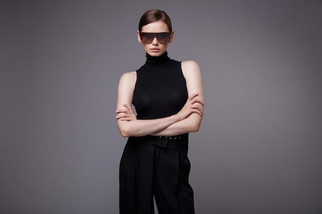 Элегантная женщина в красивых черных брюках без рукавов, стильных солнцезащитных очках позирует на сером фоне