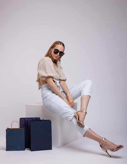elegant woman in pretty blue denim jeans, beige top, sunglasses, shopping bags on white background
