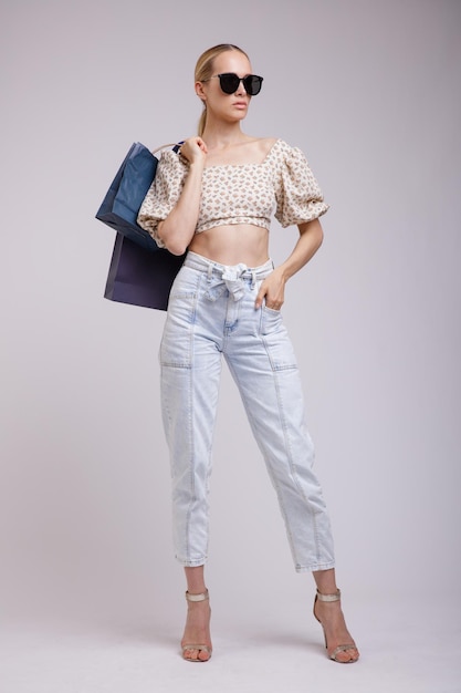 elegant woman in pretty blue denim jeans, beige top, sunglasses, shopping bags on white background