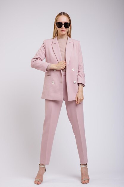 elegant woman in pretty pastel pale pink suit, jacket, trousers, sunglasses on white background