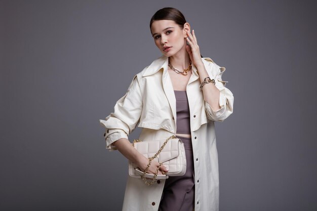 Elegant woman in pretty white coat  purple top pants  accessories handbag on gray background