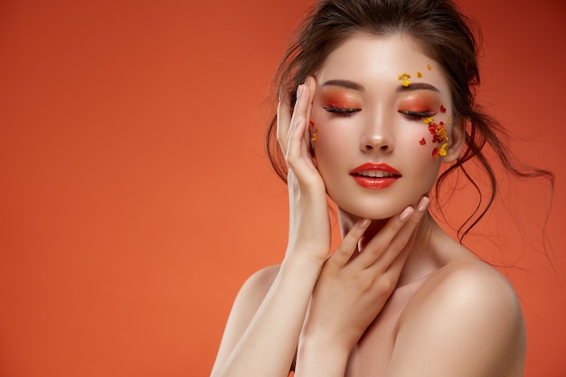 Photo elegant woman with orange make-up anf natural flowers touching her face on orange background