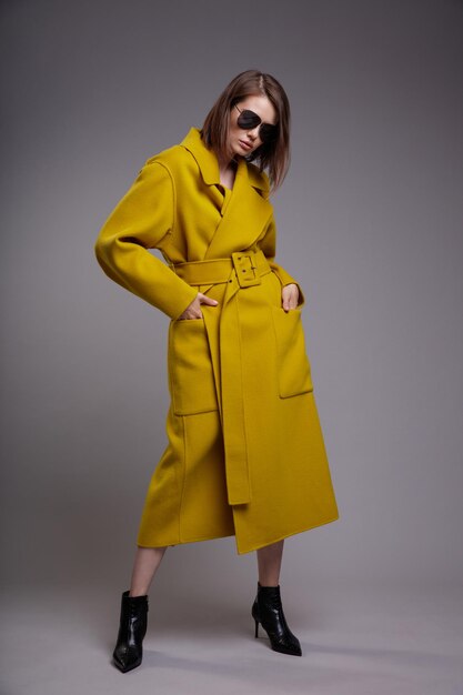 Elegant woman in yellow mustard coat black ankle boots sunglasses gray background Bob haircut