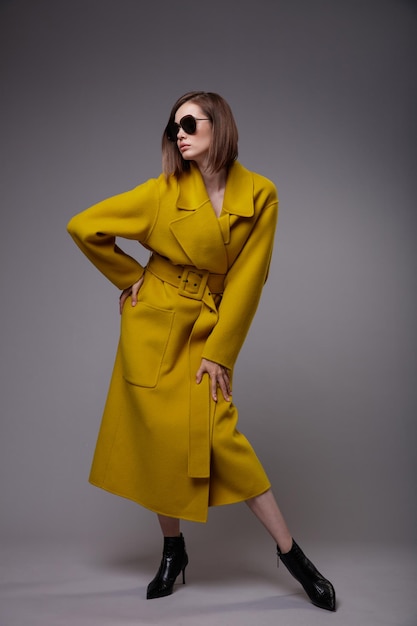 Элегантная женщина в желтом горчичном пальто, черные ботильоны, солнцезащитные очки, серый фон, стрижка Боб