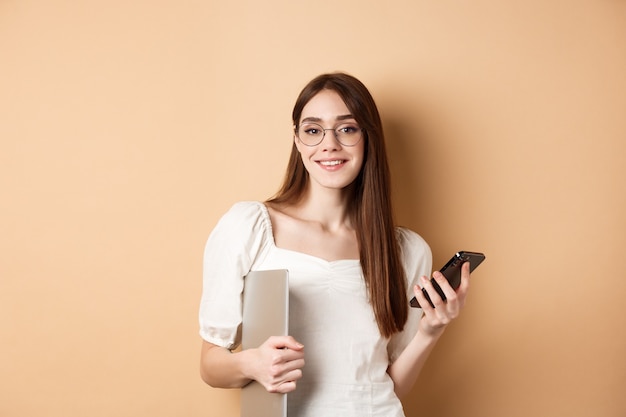 Элегантная работающая женщина в очках, держащая портативный компьютер и смартфон, довольная улыбка на камеру, стоя на бежевом фоне.