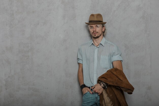 Elegant young bearded hipster man in a vintage brown hat with a brown fashionable jacket in a classic shirt posing in a room near a gray wall.