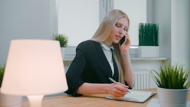 Elegant young blond female in office suit sitting at workplace with computer and negotiating via mob...