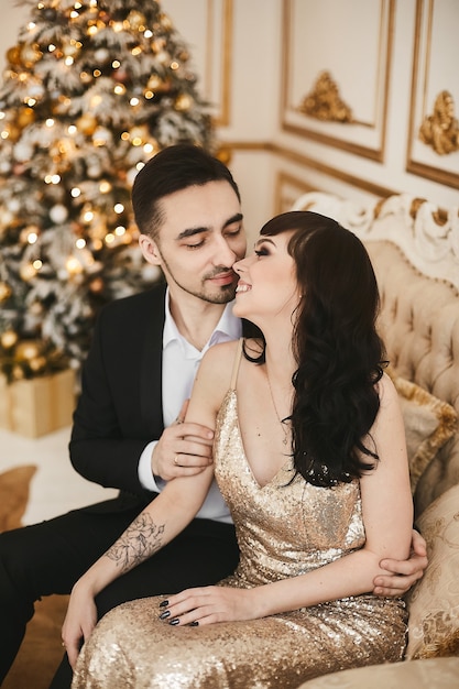 An elegant young couple in luxurious outfits sits next to a christmas tree new year party