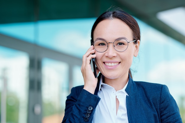 Elegant young female employee with toothy smile holding smartphone by ear while talking to one of colleagues outdoors