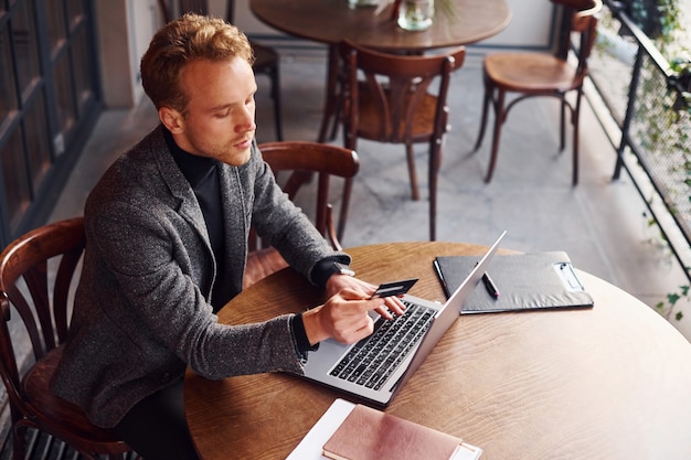 Elegante giovane ragazzo in abbigliamento formale si siede in un caffè con il suo computer portatile e la carta di credito in mano.