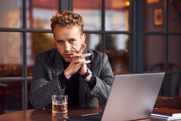 Elegant young guy in formal wear sits in cafe with his laptop and glass of alcohol.