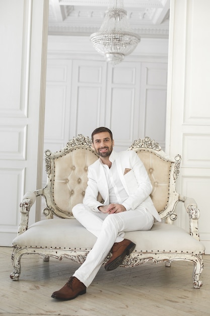 Elegant young handsome man with a beard in a white classic suit. The hotel's interior. Photo Studio. Sofa. Window. Chair. Chandelier