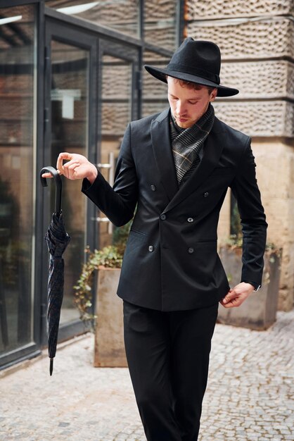 Elegant young man in black hat and clothes with umbrella in hands outdoors in the city