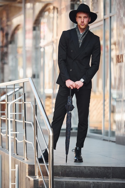 Элегантный молодой человек в черной шляпе и с зонтиком на улице в городе
