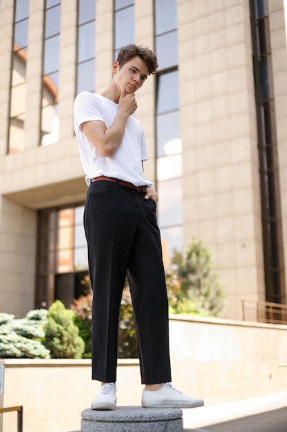 Elegant young man in a fashionable black shirt in a white stylish t-shirt in black pants with a trendy hairstyle rests near a modern business center. Attractive guy in the street on a summer day.