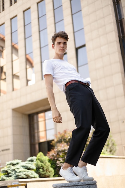 Il giovane elegante in una camicia nera alla moda in una t-shirt bianca alla moda in pantaloni neri con un'acconciatura alla moda riposa vicino a un moderno centro d'affari. ragazzo attraente in strada in una giornata estiva.