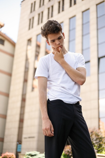 Элегантный молодой человек в модной черной рубашке в стильной белой футболке и черных брюках с модной прической отдыхает возле современного бизнес-центра. Привлекательный парень на улице в летний день.