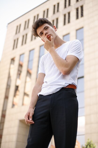 Elegant young man in a fashionable black shirt in a white stylish t-shirt in black pants with a trendy hairstyle rests near a modern business center. Attractive guy in the street on a summer day.