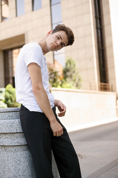 Elegant young man in a fashionable black shirt in a white stylish t-shirt in black pants with a trendy hairstyle rests near a modern business center. Attractive guy in the street on a summer day.