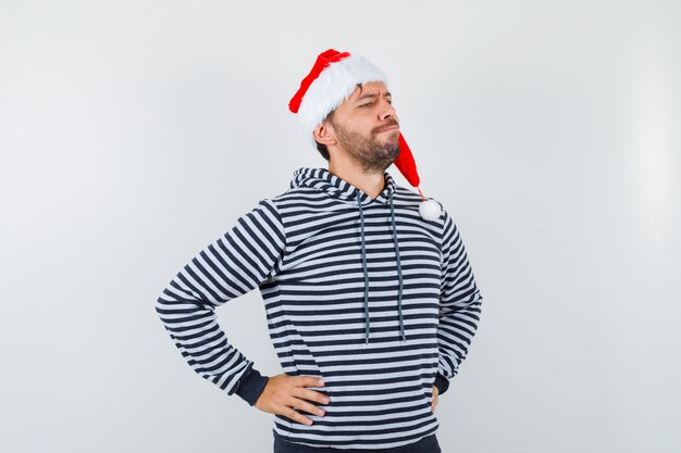 Elegant young man keeping hands on waist, looking away in hoodie, Santa hat and looking thoughtful ,