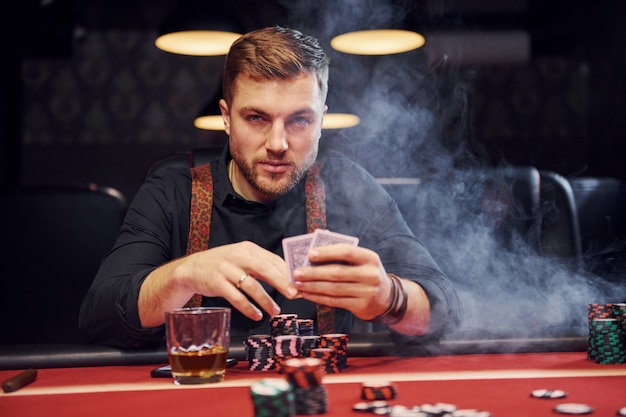 Elegant young man sits in casino with smoke in it and plays poker game