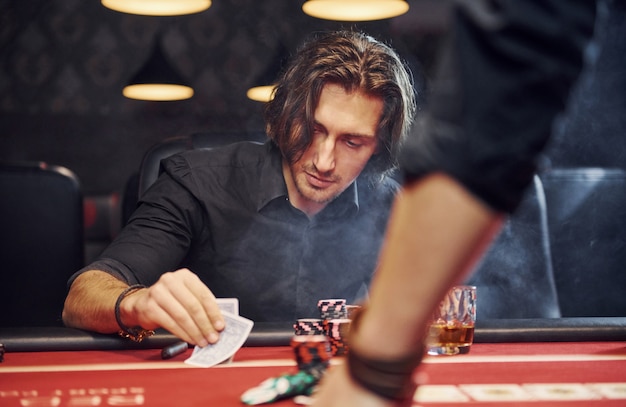 Elegant young people sits by table and playing poker in casino with smoke in the air